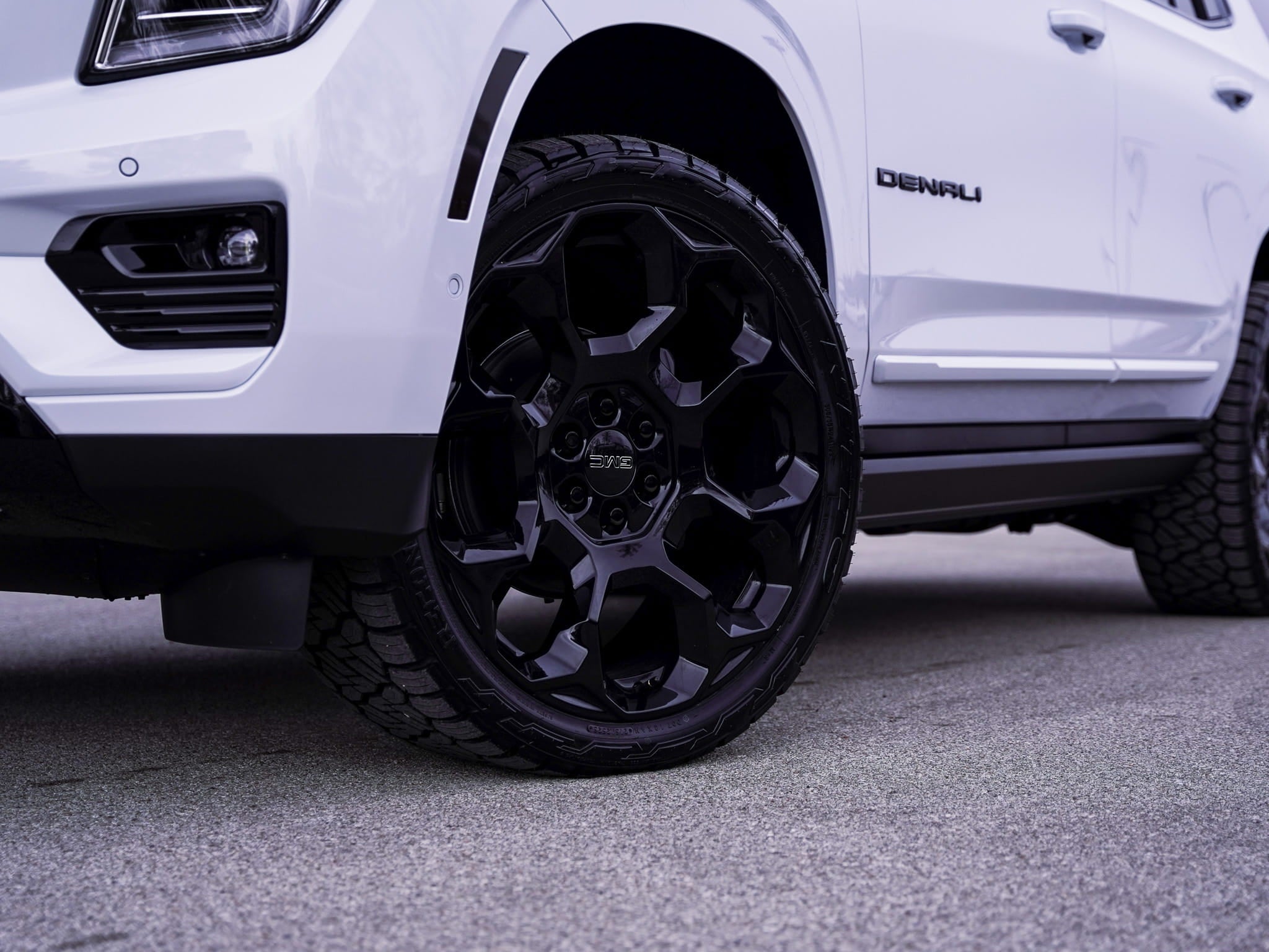 Close-up of the black powder-coated 24-inch wheel and NITTO Recon Grappler tire on a custom 2026 GMC Yukon Denali in Glacier White Tricoat at Dave Sinclair Buick GMC